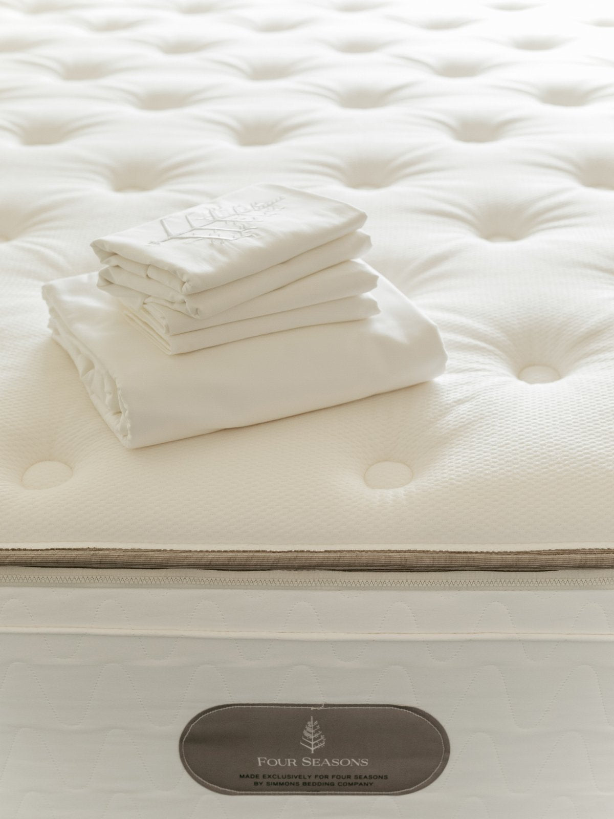 Stack of Signature White linens with logo store embroidery on a tufted mattress; border label also shows logo store. {{Signature White}}