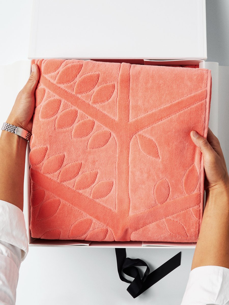 A pair of hands holding a folded beach towel with a FourSeasons logo design. The towel rests in a white box with a black ribbon. - {{Red Sea Coral}}