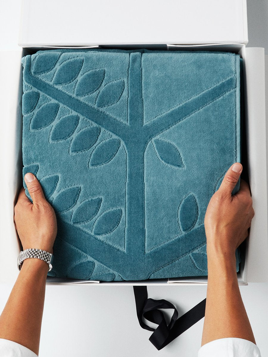 A pair of hands holding a folded beach towel with a FourSeasons logo design. The towel rests in a white box with a black ribbon. - {{Polynesian Lagoon}}