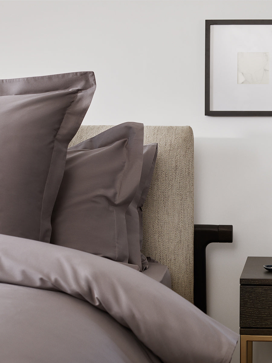 Close-up of bed with charcoal grey duvet and flange-edge pillows, textured headboard, and a dark wood nightstand with framed art on the wall. {{Hampshire Charcoal}}