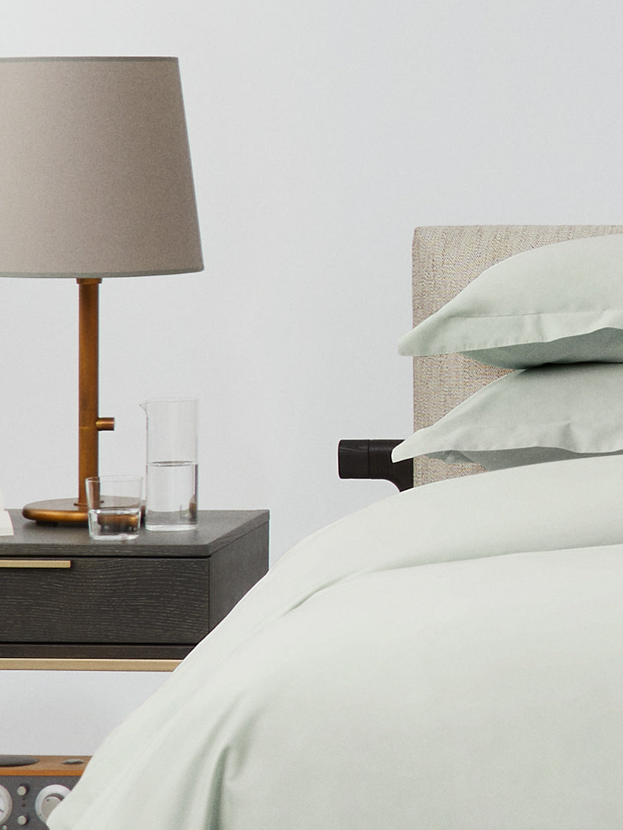 Close-up of bed with Florentine Sage bedding; stacked pillows and duvet beside a dark wood nightstand with beige lamp, glass, and carafe. {{Florentine Sage}}