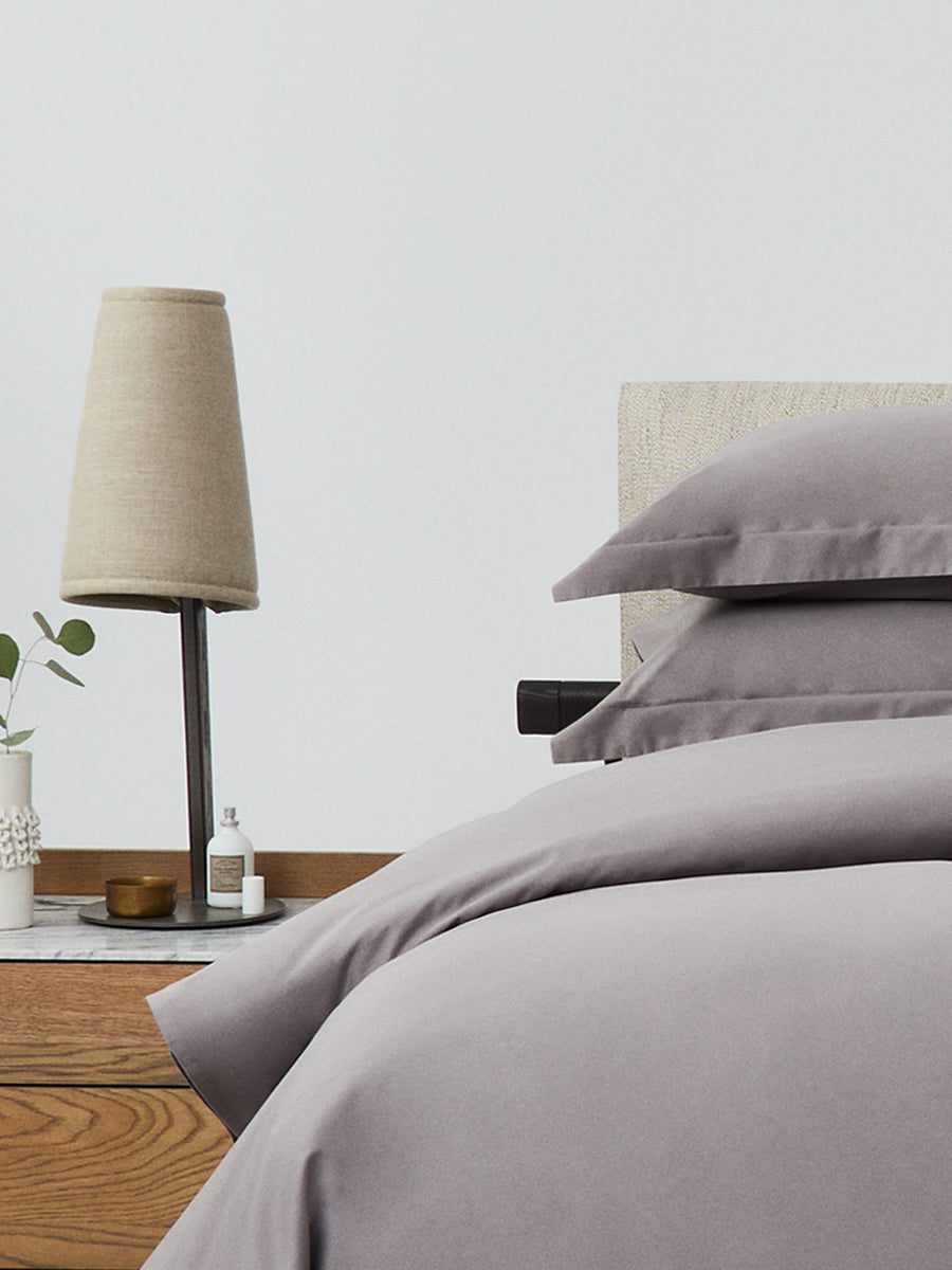 Close-up of bed with Hampshire Charcoal bedding; stacked pillows and duvet beside a wood nightstand with beige lamp and decor. {{Hampshire Charcoal}}