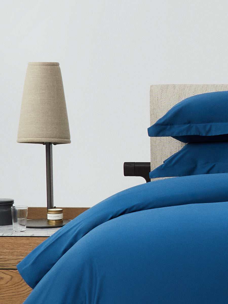 Close-up of bed with Riviera Navy bedding; stacked pillows and duvet beside a wood nightstand with beige lamp, candle, and glass. {{Riviera Navy}}