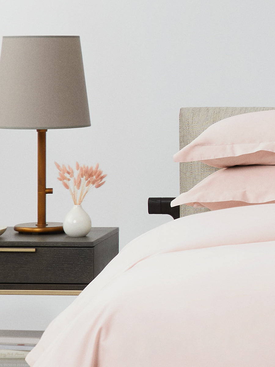 Close-up of bed with Kyoto Blush bedding; stacked pillows and duvet beside a dark wood nightstand with beige lamp and small vase. {{Kyoto Blush}}