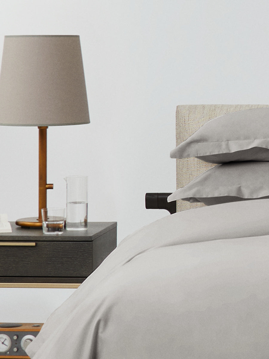Close-up of bed dressed in Spanish Slate bedding; stacked pillows and duvet, bedside table with taupe lamp, glass and carafe. {{Spanish Slate}}