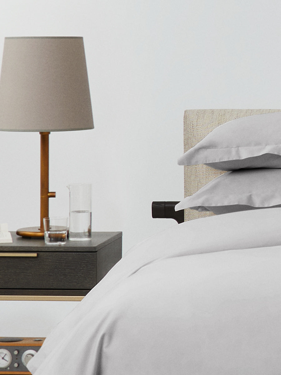 Side view of Turkish Silver duvet with stacked pillow shams beside a dark wood nightstand with lamp, glass, and carafe.{{Turkish Silver}}