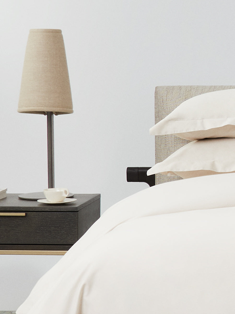 Close-up of bed with Balinese Pearl bedding; stacked pillows and duvet beside a dark wood nightstand with beige lamp and cup. {{Balinese Pearl}}