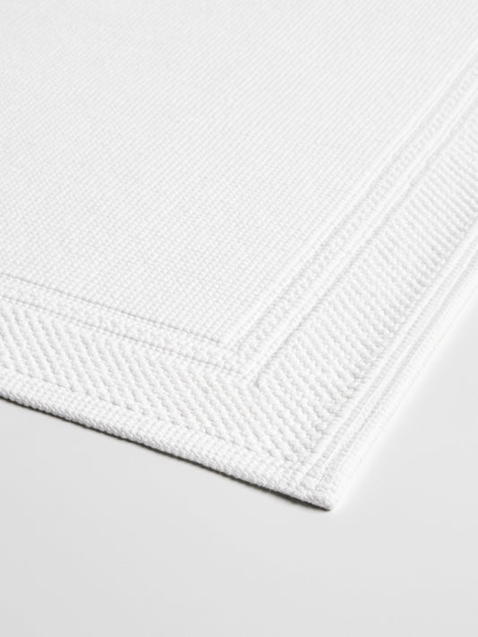 Close-up of a white bath mat with a textured surface and a subtle border. {{White}}
