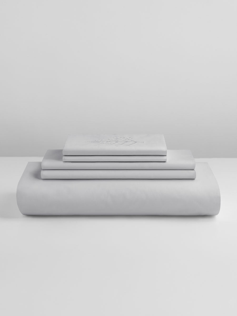 Stacked Turkish Silver duvet cover set with folded shams and pillowcases, top piece featuring store logo embroidery on a light grey background. {{Turkish Silver}}