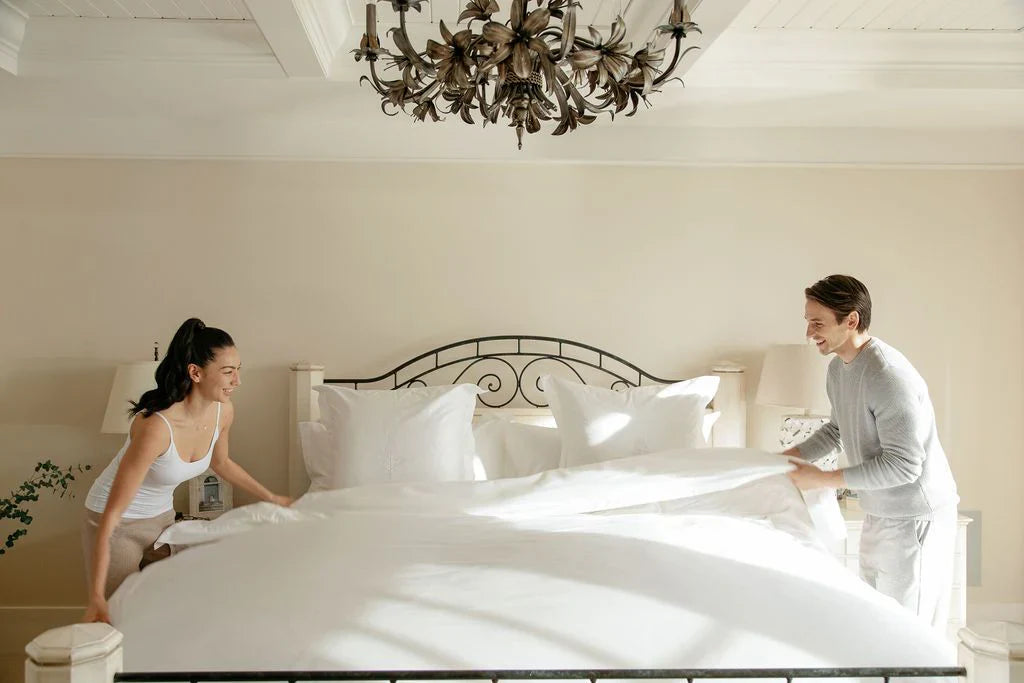 Smiling couple making a bed together in a bright, elegant bedroom with white bedding and a decorative chandelier overhead.
