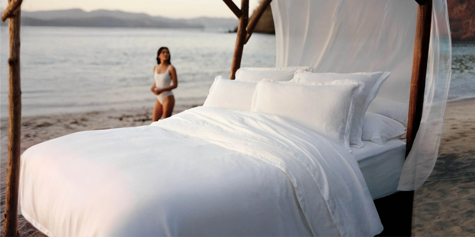 Luxurious white bed with plush pillows and flowing canopy set on a beach, with a woman in a white swimsuit standing near the shoreline.