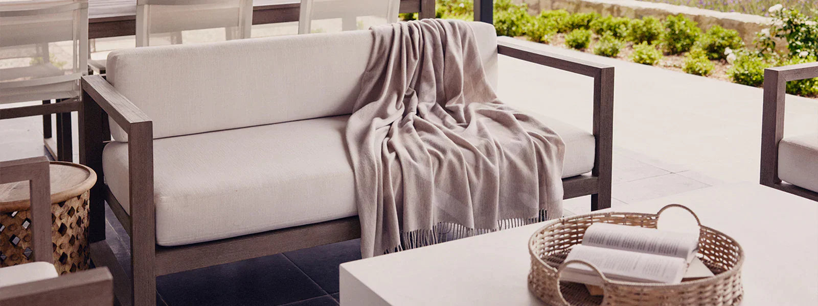 Outdoor patio sofa with a beige throw blanket draped over the armrest, next to a white table with a woven tray and open book.
