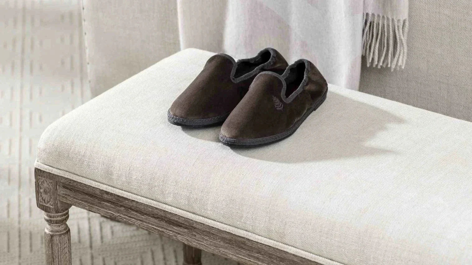 Pair of dark brown velvet slippers placed on a light upholstered bench with a fringed throw in the background.