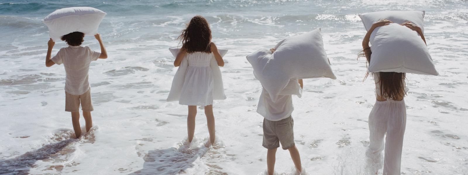 Four people standing at the edge of the ocean, each holding a white pillow as the waves gently surround their feet.