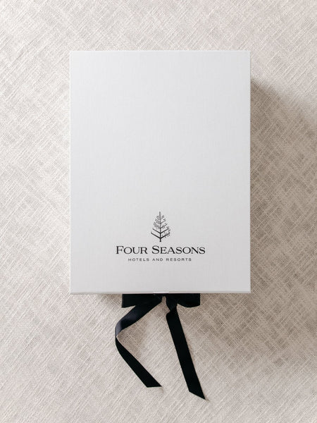 A white box with a black ribbon tied around it. The Four Seasons Hotels and Resorts logo is printed on the bottom of the box.