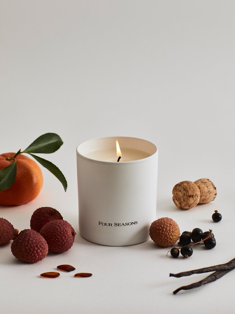 A lit Four Seasons candle from the Candle Set,  surrounded by ingredients like lychees and blackcurrants