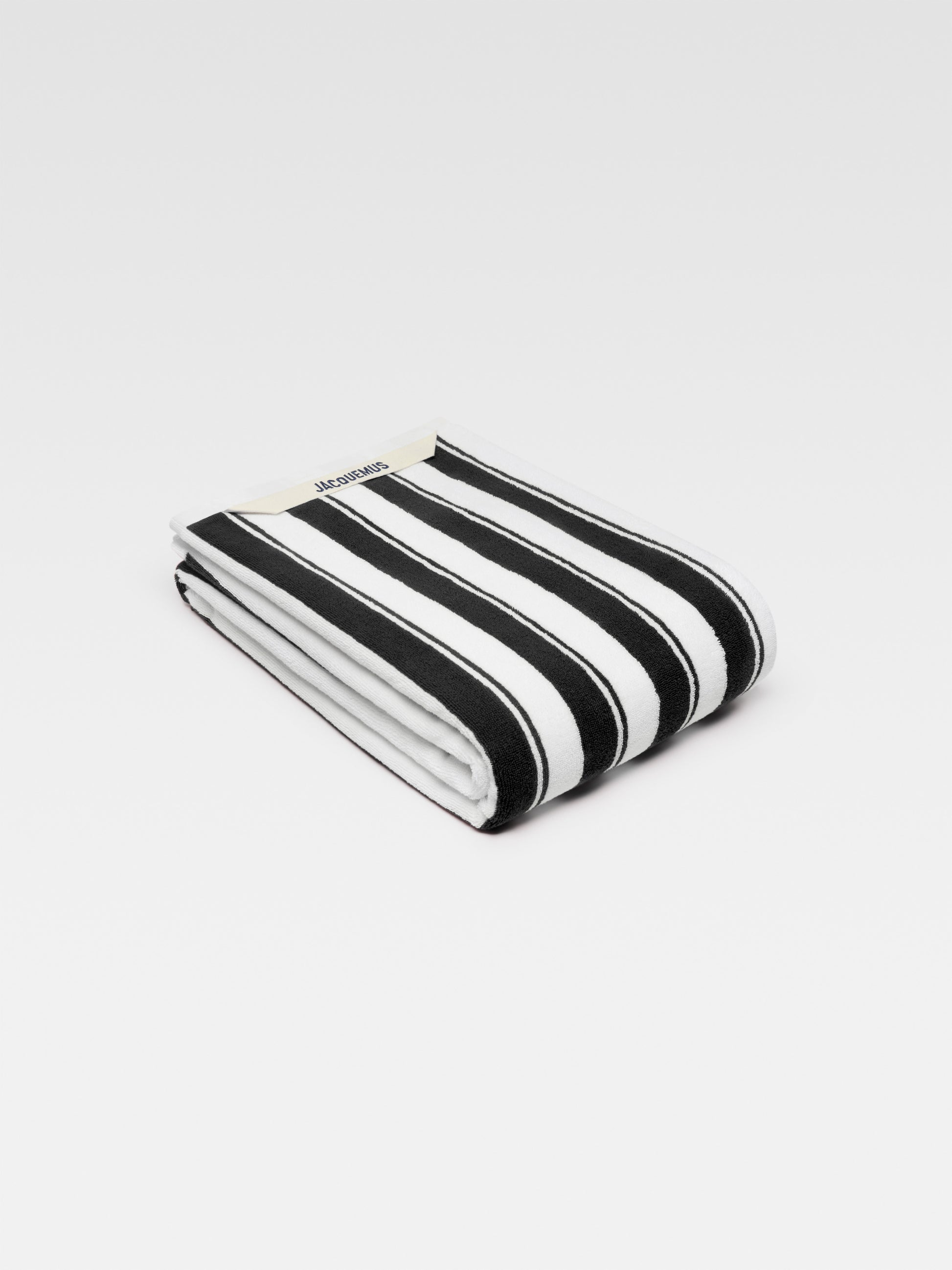 A large, folded beach towel with bold black and white stripes. The Jacquemus logo is visible in the top left corner.