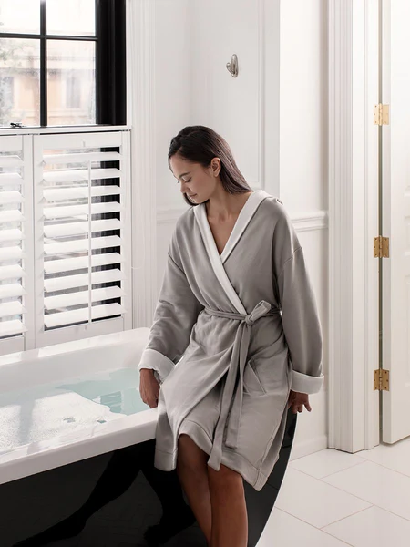 Woman in a gray bathrobe sitting on the edge of a bathtub in a bright, elegant bathroom.
