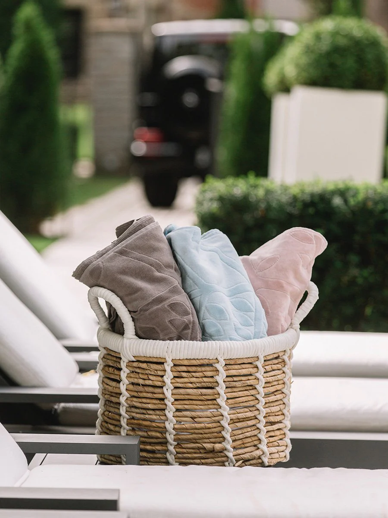 Wicker basket with folded towels in gray, blue, and pink by a row of poolside lounge chairs.