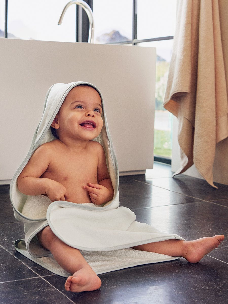 A baby wearing a hooded towel, smiling while sitting near a bathtub. - {{Coral Reef Pink / Mountain Grey}}