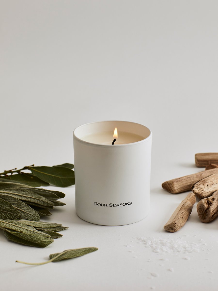 A lit Four Seasons candle from the Candle Set,  styled with coastal elements like sage leaves and driftwood.