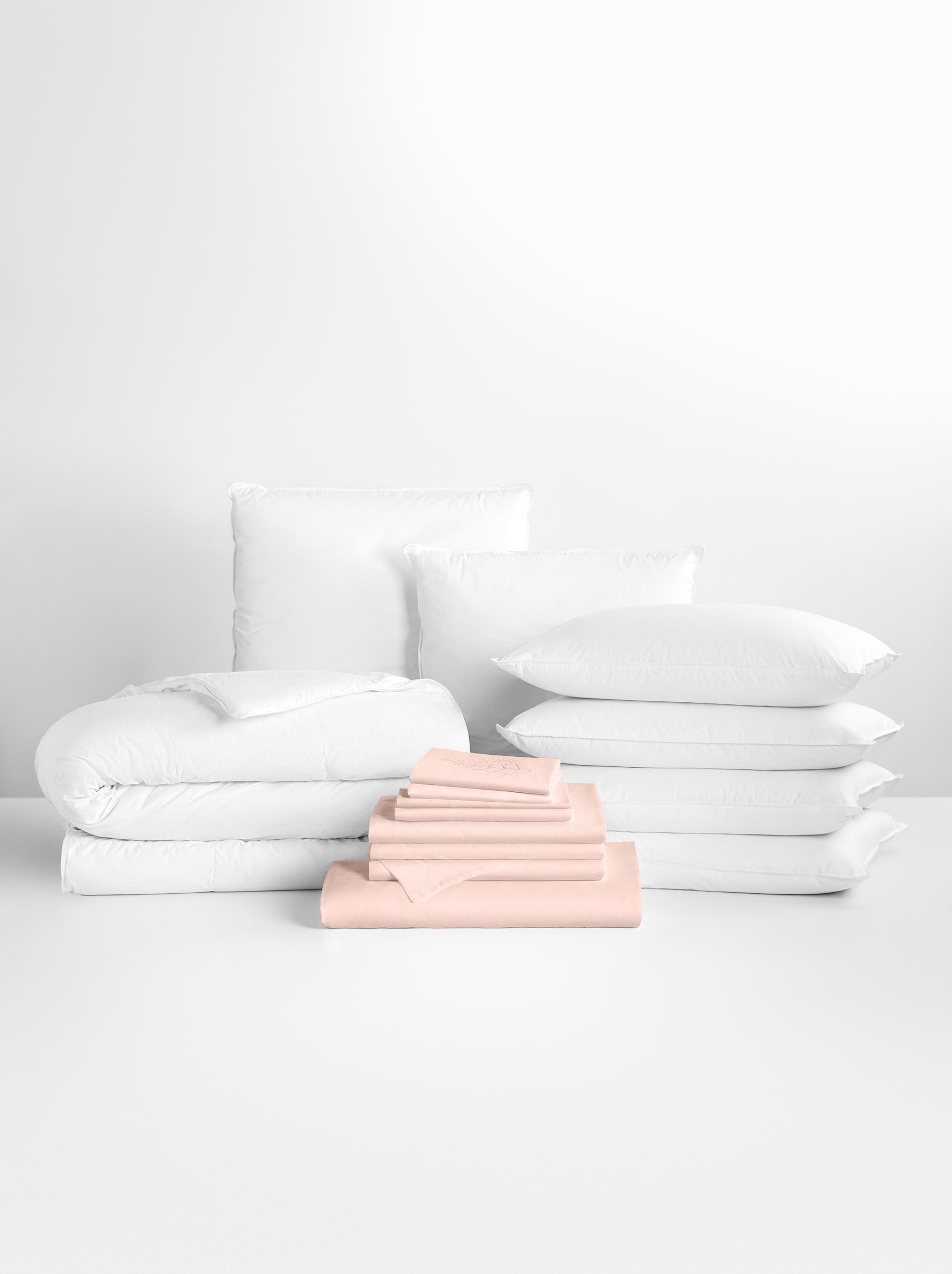 Kyoto Blush-colored sheet set in the foreground, with stacked pillows and a neatly folded duvet in the background.