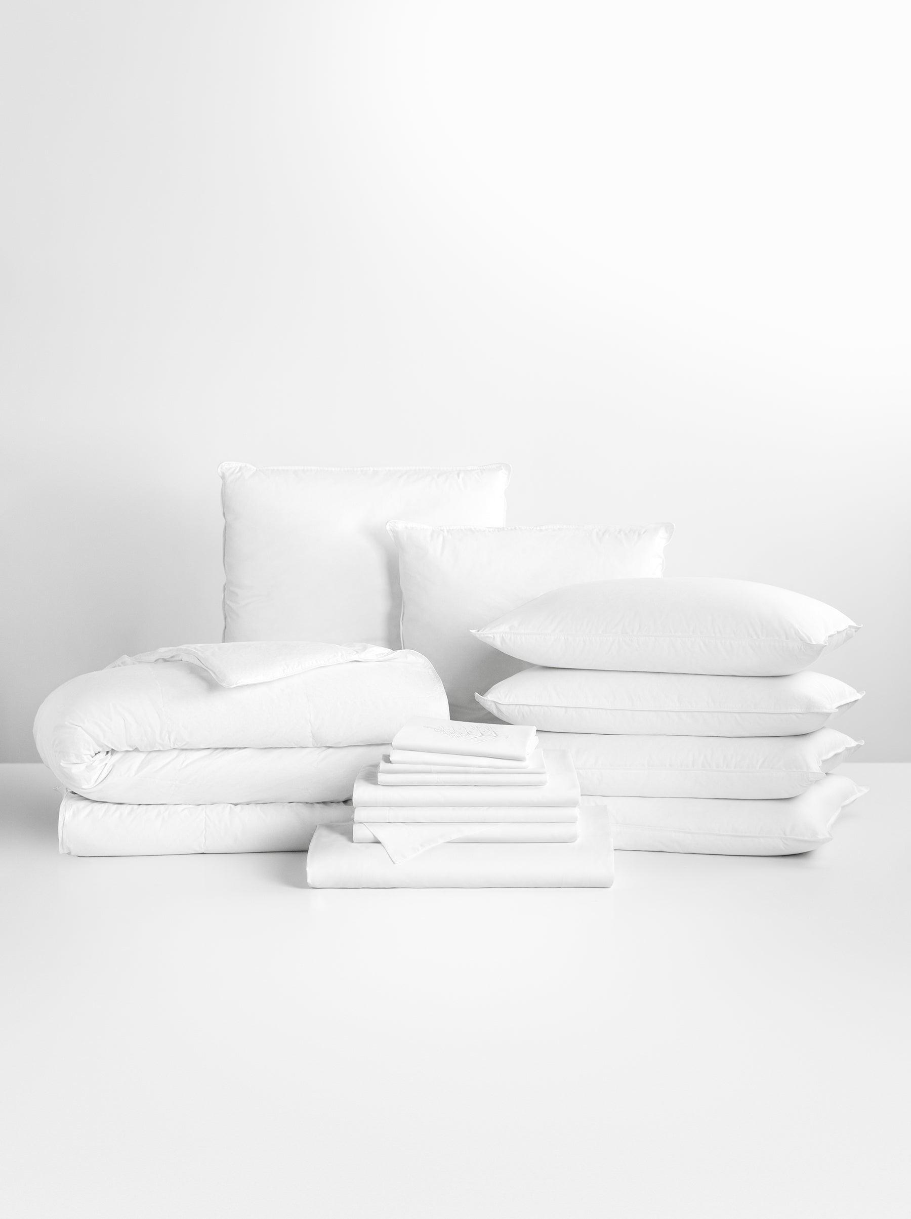 Complete bedding set in white tones. Includes a voluminous duvet, several pillows of different sizes and a stack of plain sheets.