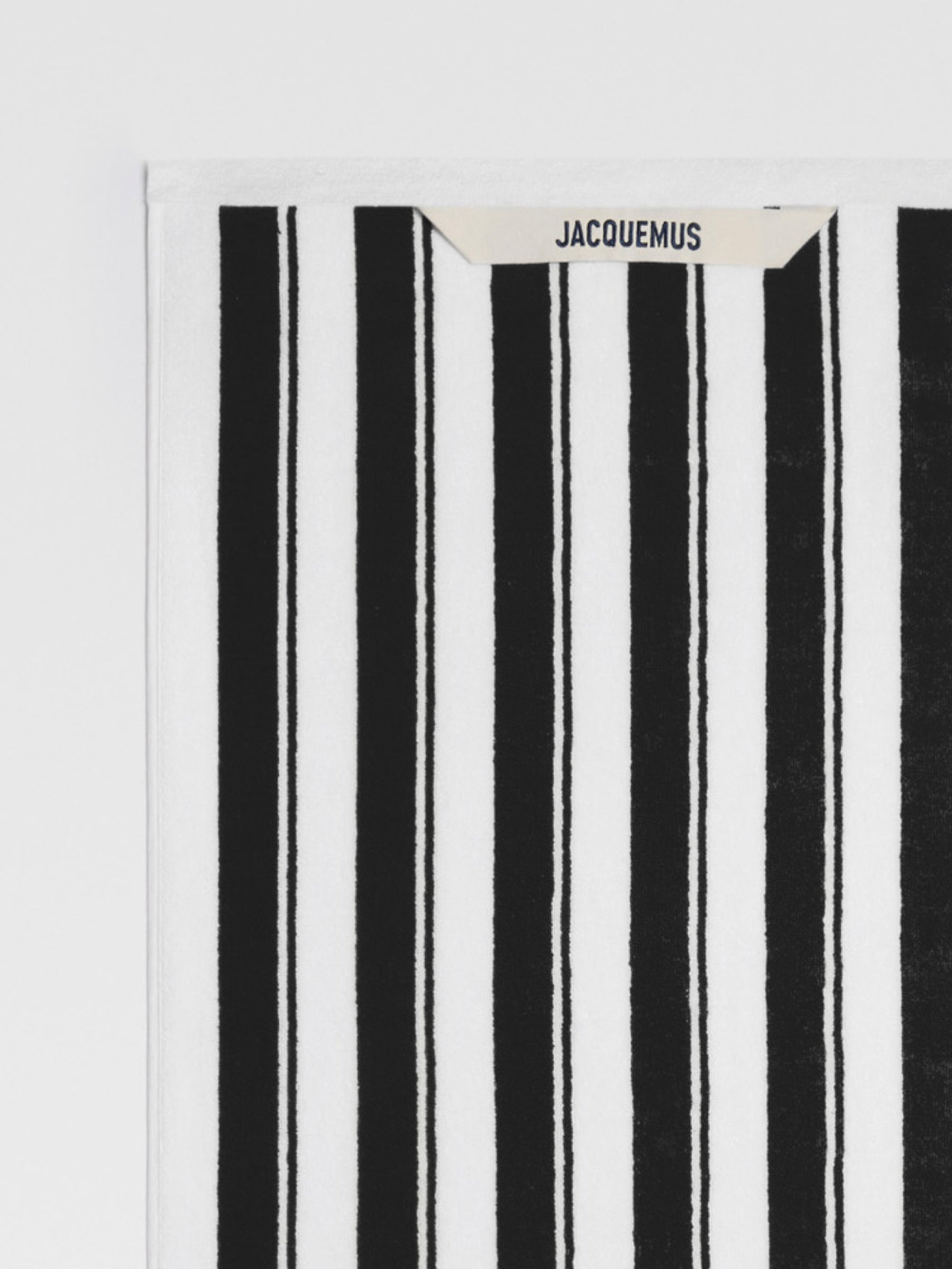 A close-up of a white beach towel with bold black stripes. The Jacquemus logo is visible in the top left corner.