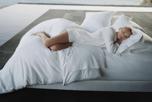 Woman in white loungewear lying on a bed with white linens beside a calm water pool, resting her head on a pillow and gazing peacefully.