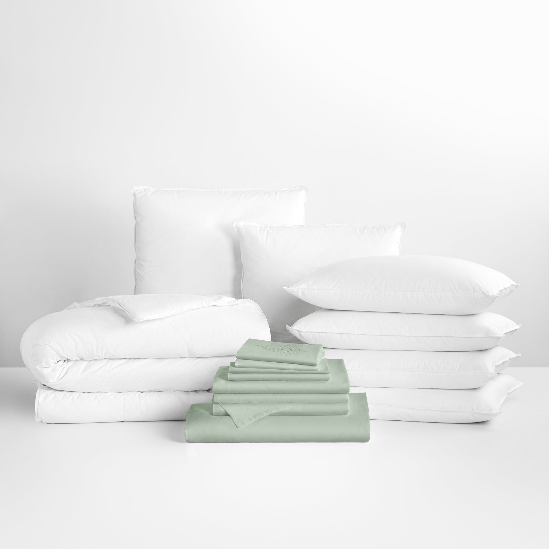Florentine Sage-colored sheet set in the foreground, with stacked pillows and a neatly folded duvet in the background. - {{Florentine Sage}}