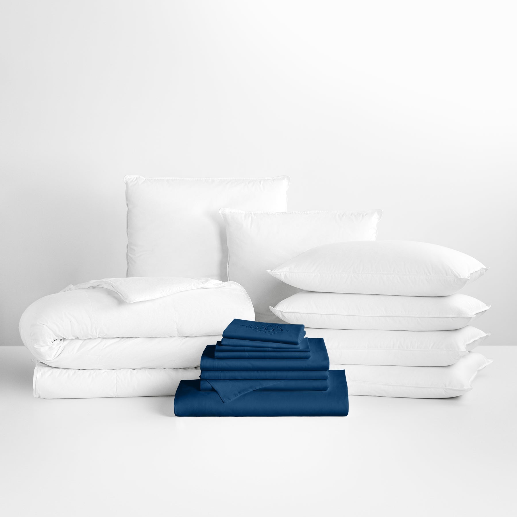 Riviera Navy-colored sheet set in the foreground, with stacked pillows and a neatly folded duvet in the background. - {{Rivera Navy}}