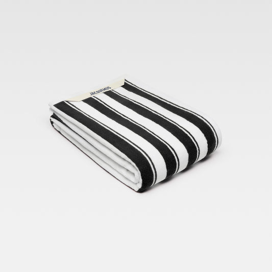 A large, folded beach towel with bold black and white stripes. The Jacquemus logo is visible in the top left corner.