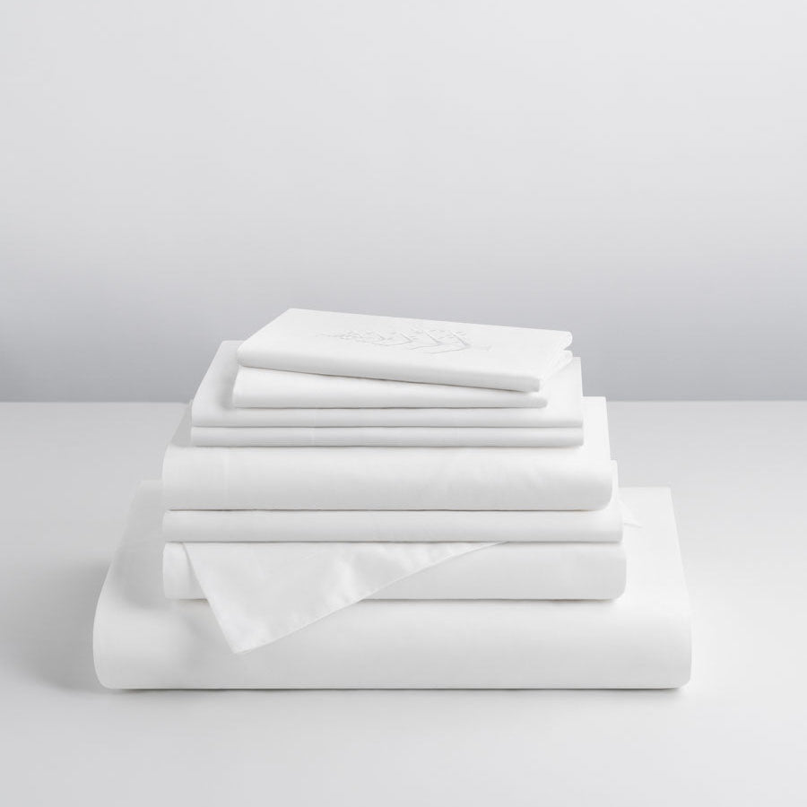 Signature Hotel Linen Set in Signature White stacked on a table—Supima cotton fitted sheet, flat sheet, pillowcases, duvet cover, pillow shams, and Euro shams with store logo. {{Signature White}}