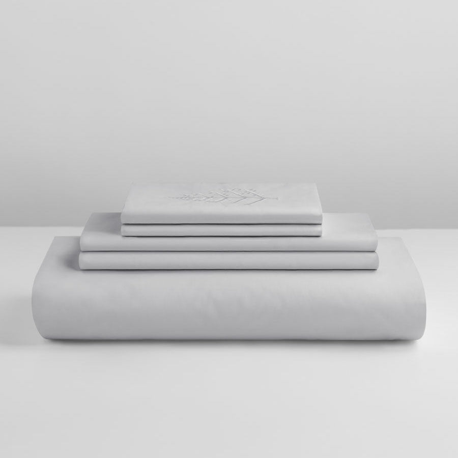 Stacked Turkish Silver duvet cover set with folded shams and pillowcases, top piece featuring store logo embroidery on a light grey background. {{Turkish Silver}}