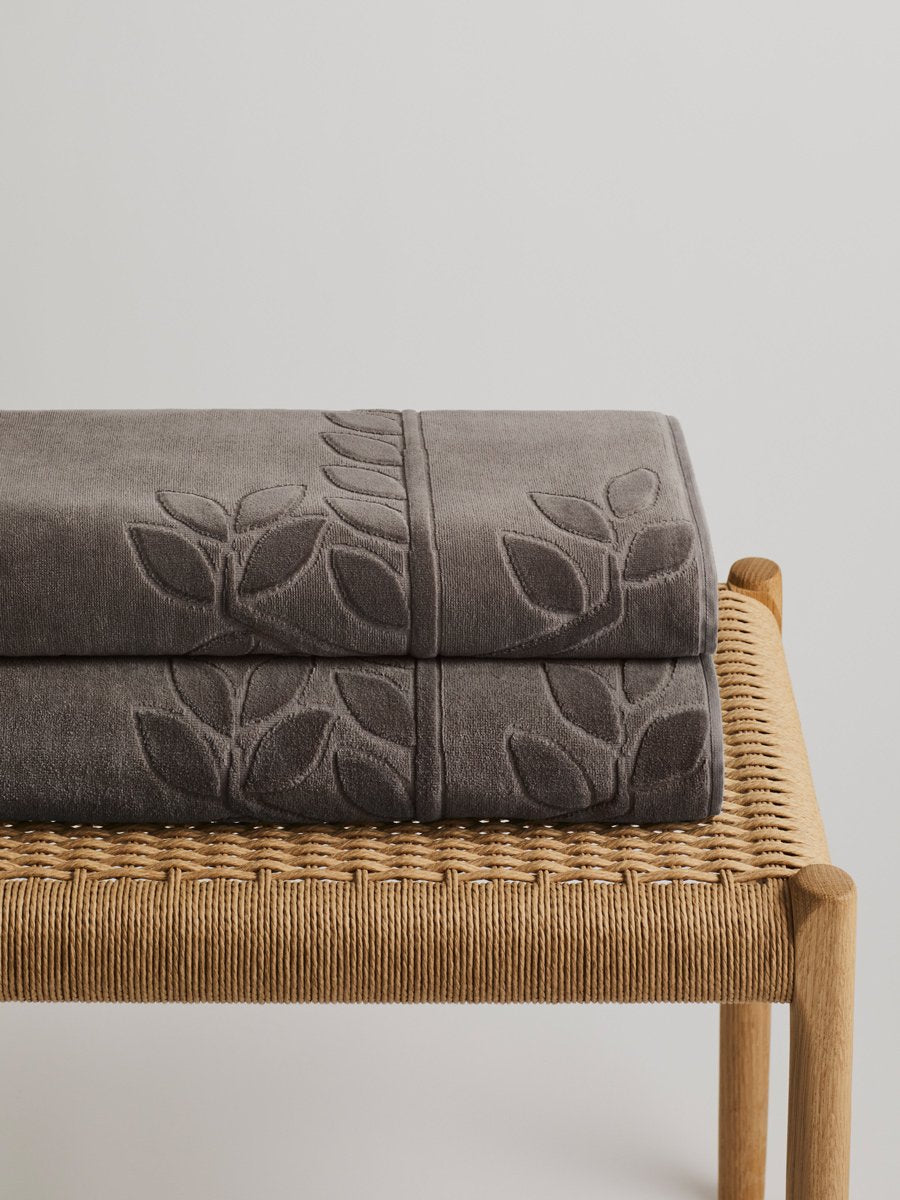 Two "Californian Driftwood" beach towels folded on a wooden bench. The towels feature a subtle leaf pattern.
