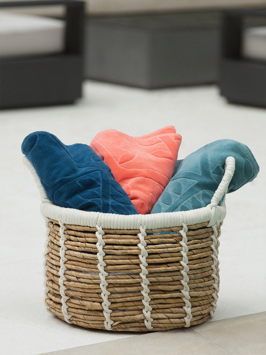 Three folded beach towels in different colors are displayed in a basket next to a poolside lounge chair.