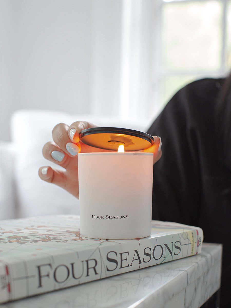 A white Four Seasons candle with a black lid featuring an embossed design.