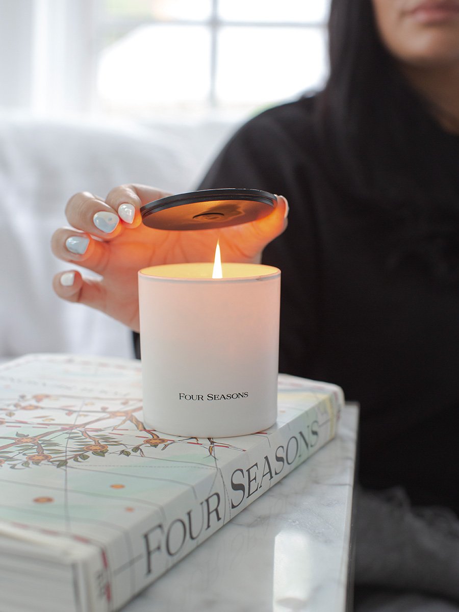 A white Four Seasons candle with a black lid featuring an embossed design.