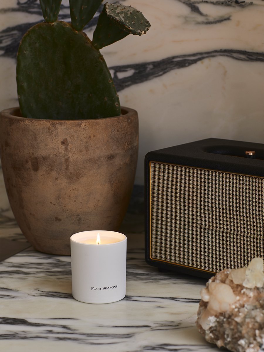 A lit Four Seasons Signature Votive - Urban Sanctuary candle creating a warm and inviting ambiance on a stylish side table.